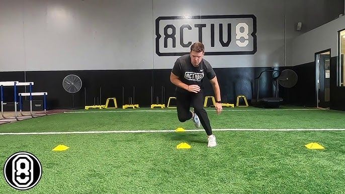 Man performing lateral agility drills with cones on an indoor turf field at a training facility.